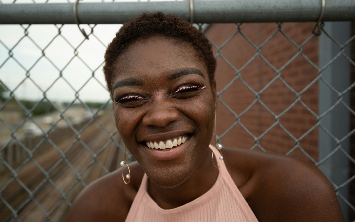 A young woman with a radiant smile, sporting short curly hair, hoop earrings, and glittery eyeliner, leans against a chain-link fence with an urban backdrop.