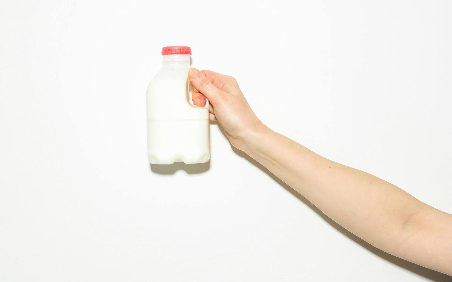 A person's arm is extended towards the camera. The person is holding a small plastic milk bottle with a red cap against a plain white background.