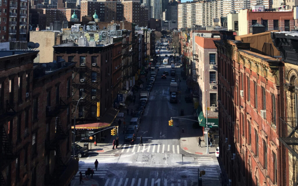 "Elevated view of a bustling city street intersection with pedestrian crosswalks, traffic lights, and rows of classic brick buildings on either side, under a clear blue sky.