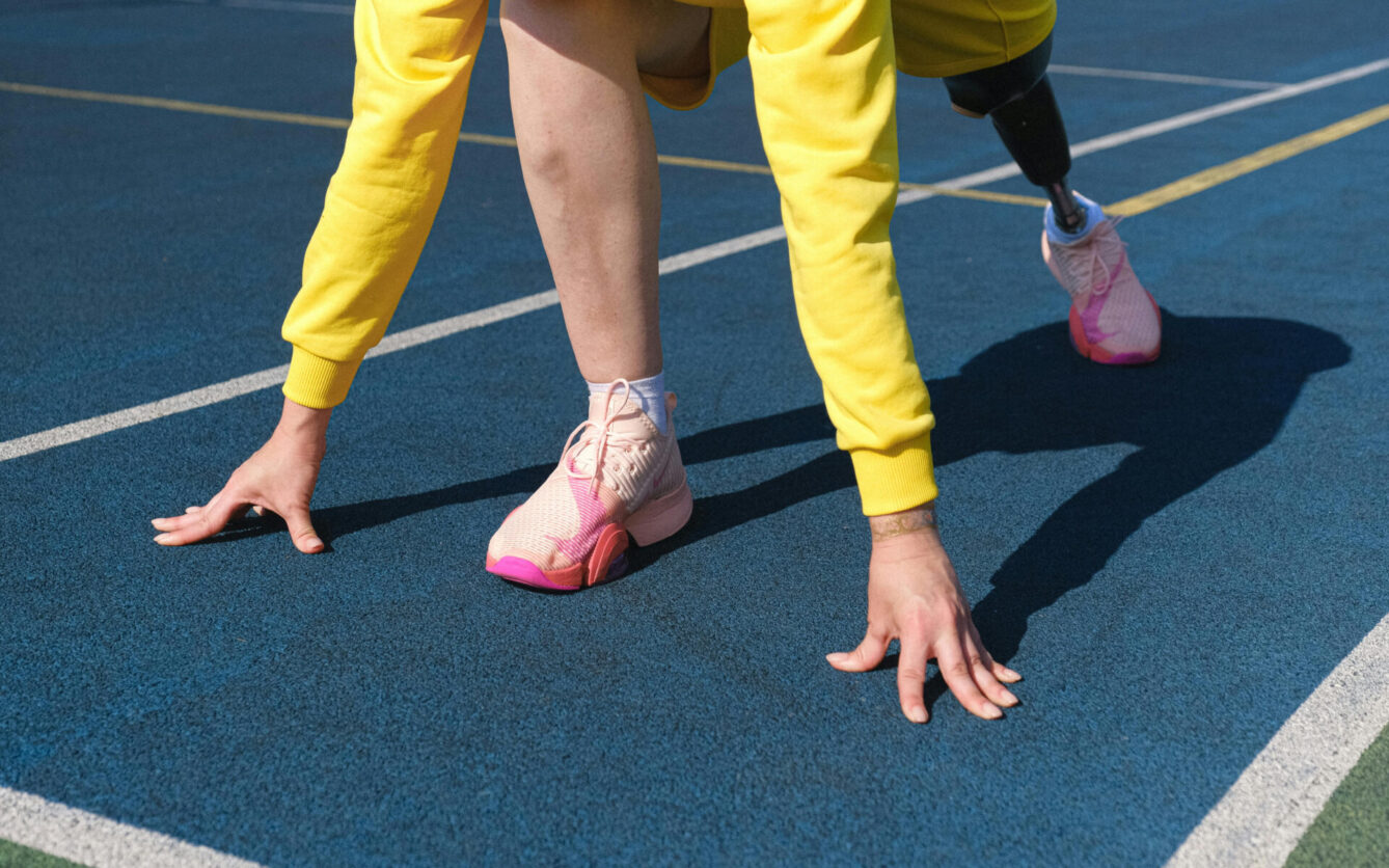 A person in a yellow sweatshirt preparing to sprint on a blue track. The individual is wearing pink athletic shoes and has a prosthetic leg, emphasizing resilience, athleticism, and inclusivity in sports.