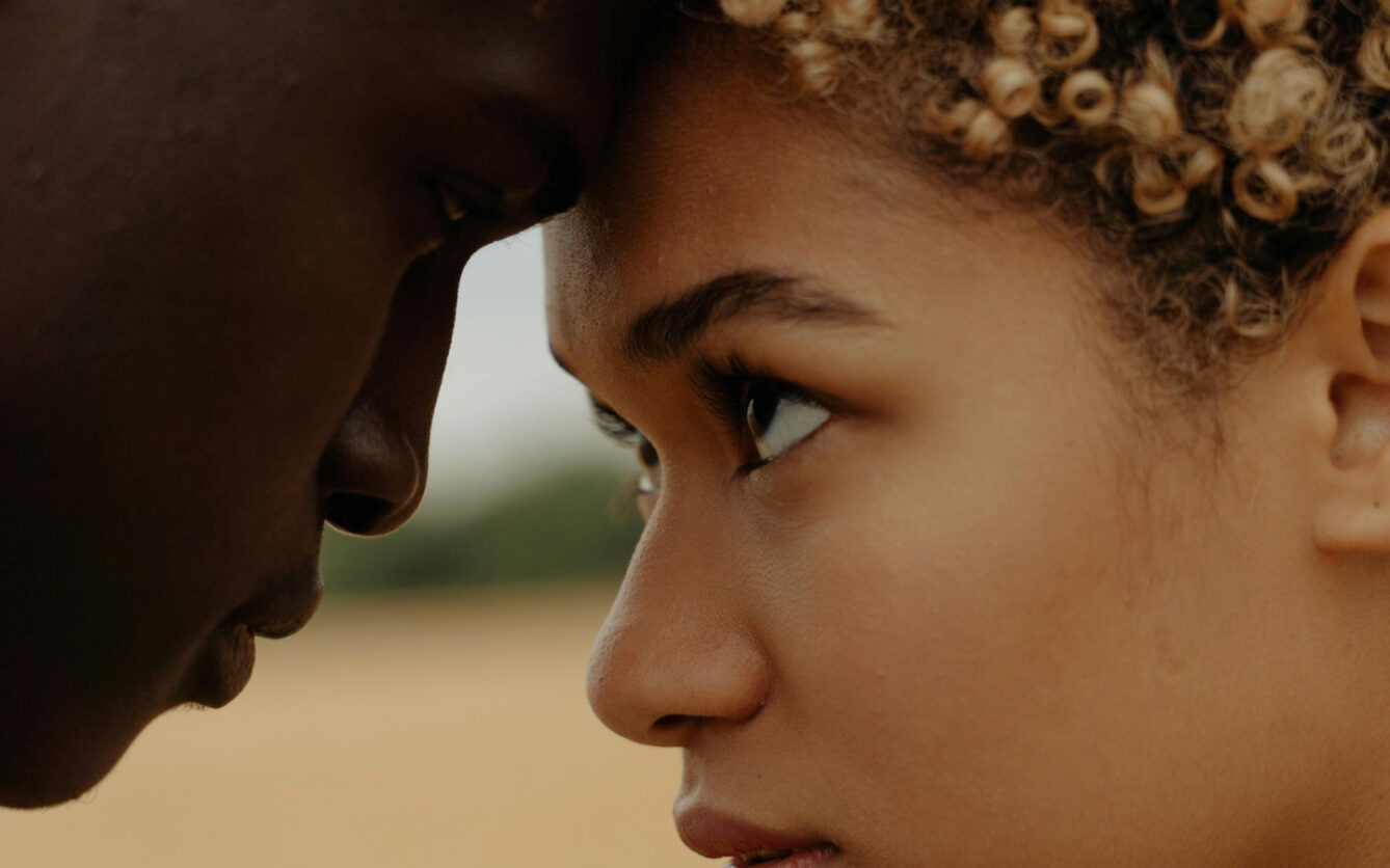 This is a close-up image of a young woman and a man touching foreheads, showcasing an intimate moment. The focus is on the woman's profile, capturing her detailed facial features and expressive eyes set against a blurred natural background.