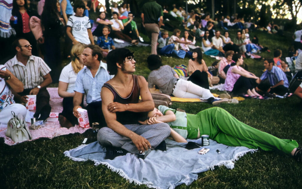 This is a vintage scene of diverse people relaxing on a grassy hill at a park during a public event. It features a man in striped pants and a tank top reclining with a camera, with others lounging and conversing around blankets spread out with snacks and drinks.