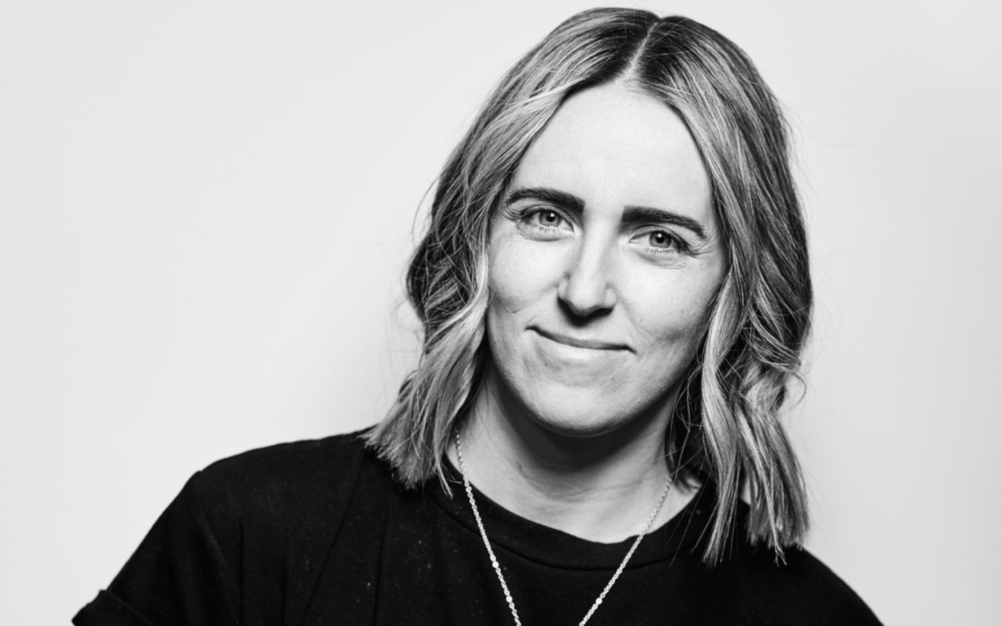 Black and white portrait of a woman with shoulder-length wavy hair, smiling gently at the camera, wearing a black top and a delicate necklace.