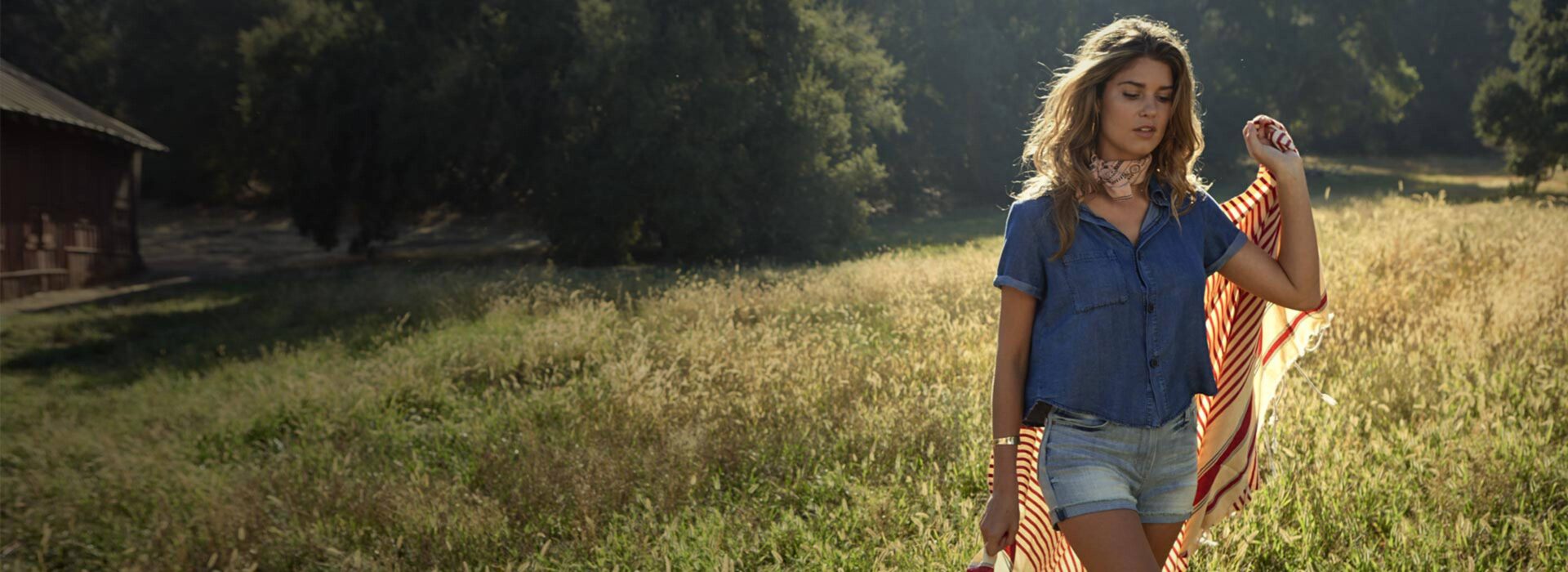 Person in denim walking through a sunlit field with a striped blanket over one shoulder