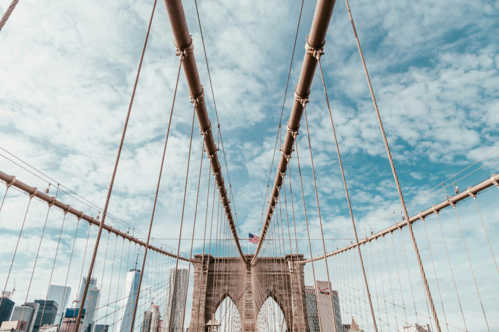 View of the Brooklyn Bridge cables and towers with an American flag against a cloudy blue sky and the New York City skyline in the background