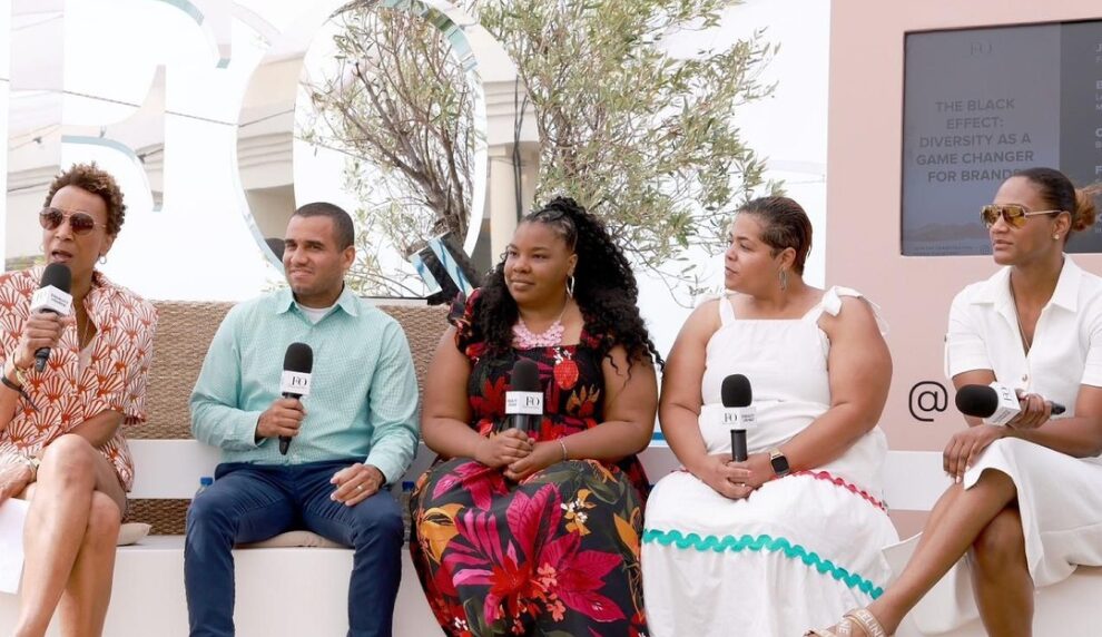 A panel of five individuals seated, holding microphones, discussing 'The Black Effect: Diversity as a Game Changer for Brands,' during an outdoor event