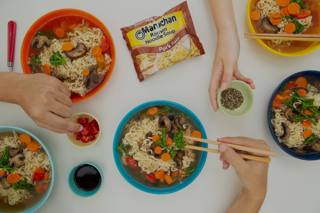 Hands preparing and serving instant ramen noodles with vegetables and pork flavor packet visible