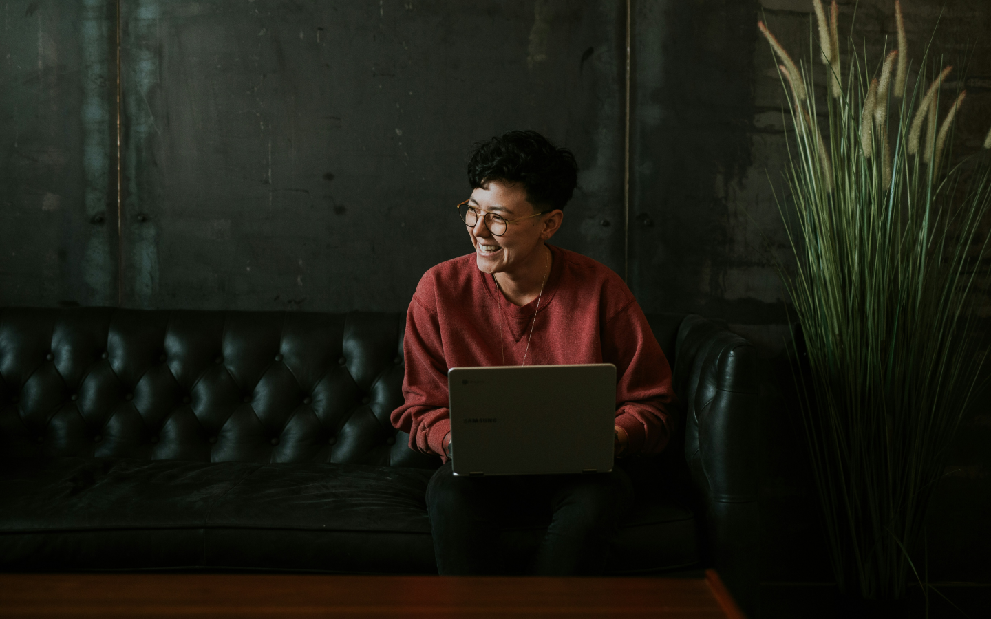 Person in a red sweater and glasses smiling and looking off to the side while sitting on a black leather couch with a laptop on their lap, in a room with a concrete wall and a large potted grass plant